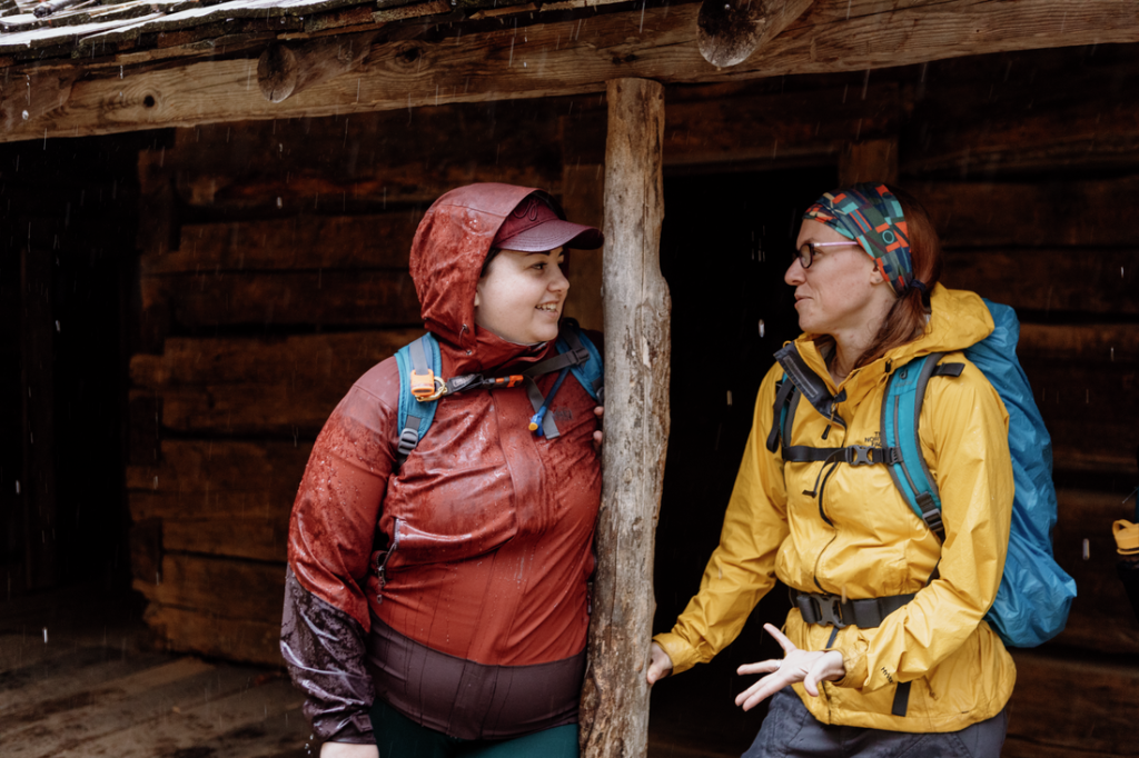 Two hikers talk in a shelter, one in a red jacket, the other in a yellow jacket.
