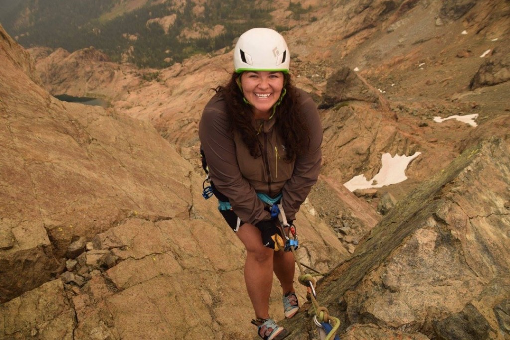 Sam Ortiz begins to rappell from an anchor on Ingalls Peak near Leavenworth, Washington. Large slabs of rock stretch down behind her to a valley far below, spotted with snow and trees.