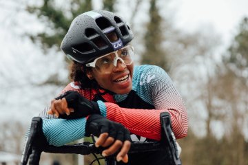 Ayesha McGowan leans on the handlebars of her bike while wearing a helmet, glasses, and a bike jersey