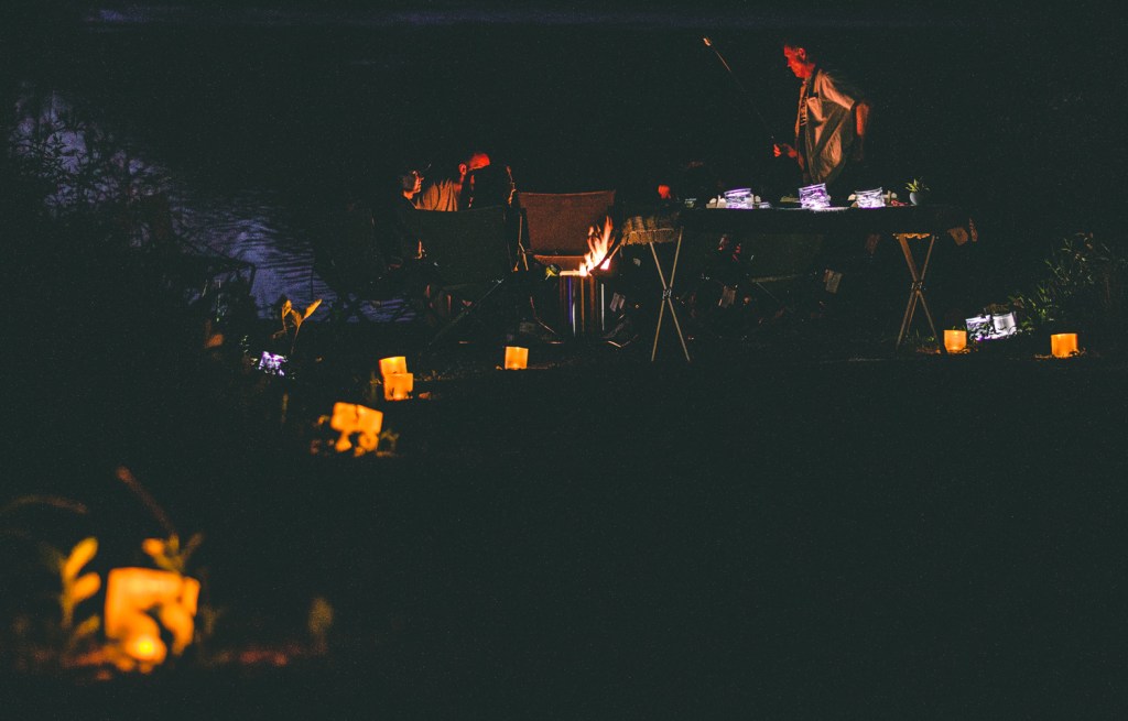 People sit around a campfire while lanterns light the night up around them.