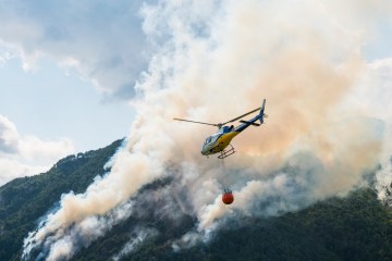 A helicopter is flying over a forest fire with a container of water beneath it.