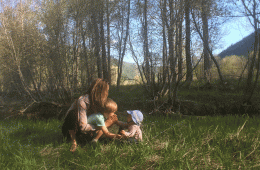 A woman and two small children practice yoga in a green field with sunlight shimmering in the background