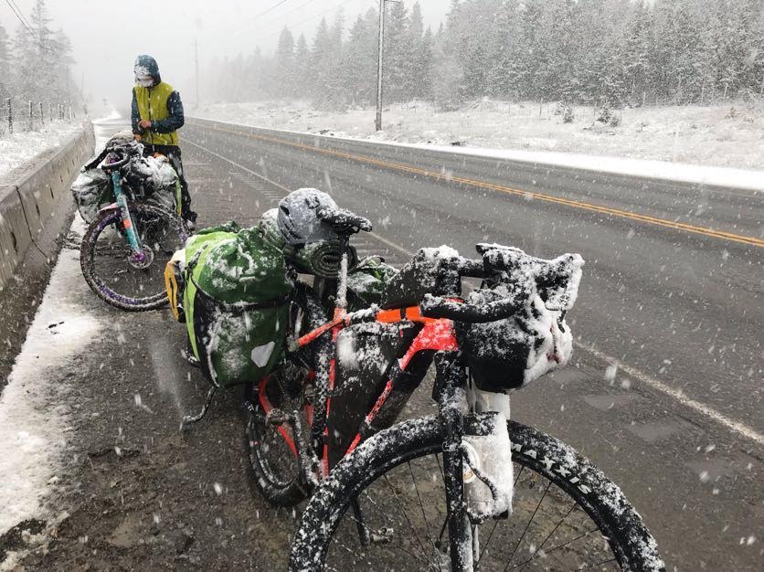 A man stands in the snow next to the road and two bicycles packed full of gear