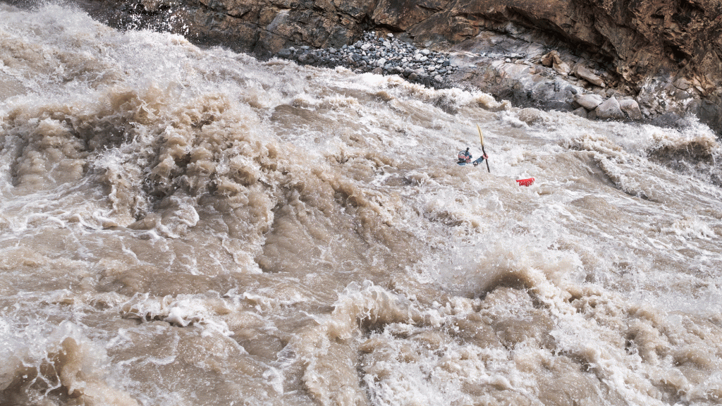 A tiny kayaker can be seen among raucous brown, splashy whitewater waves 