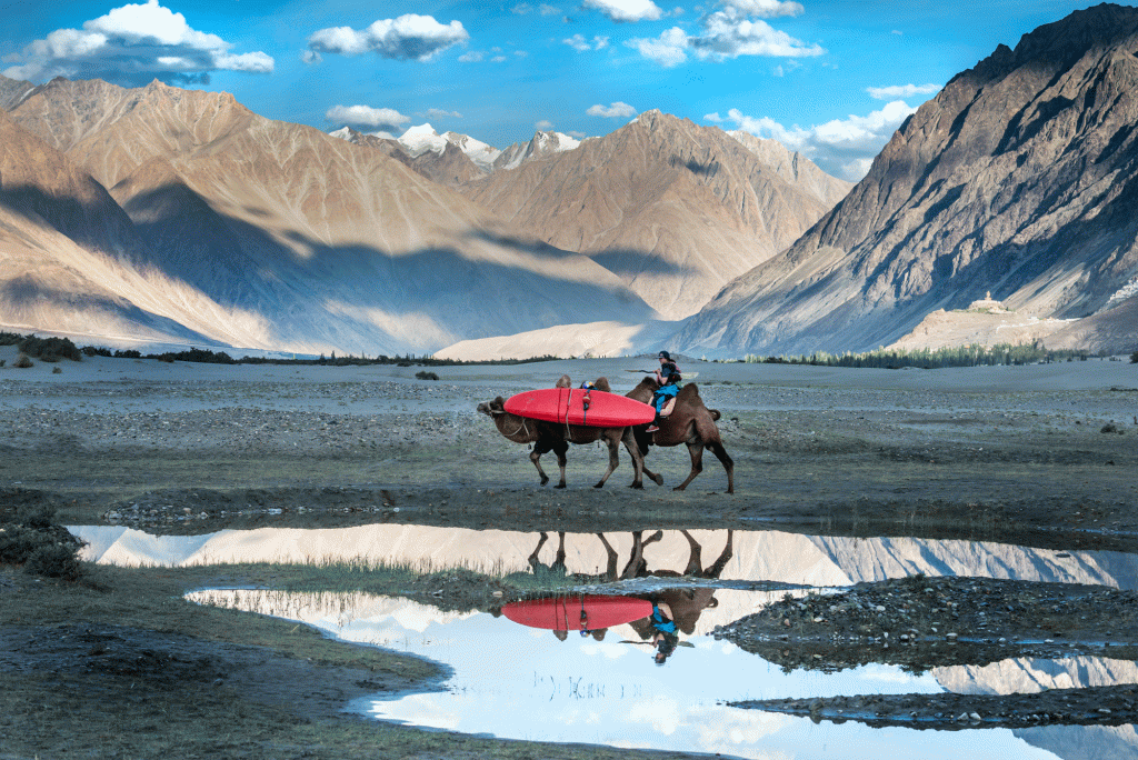 A kayaker crosses a wide-open plain on a camel with her kayak. There are mountains and bright-blue sky in the distance.