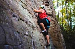 Sam Ortiz steps up to the next hold on a grey rock in the Red River Gorge, with green trees behind.