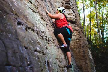 Sam Ortiz steps up to the next hold on a grey rock in the Red River Gorge, with green trees behind.