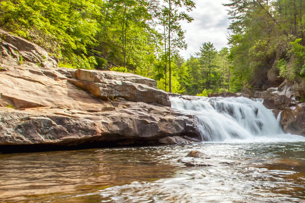 A rippling pool above light brown rocks beneath a rushing waterfall.