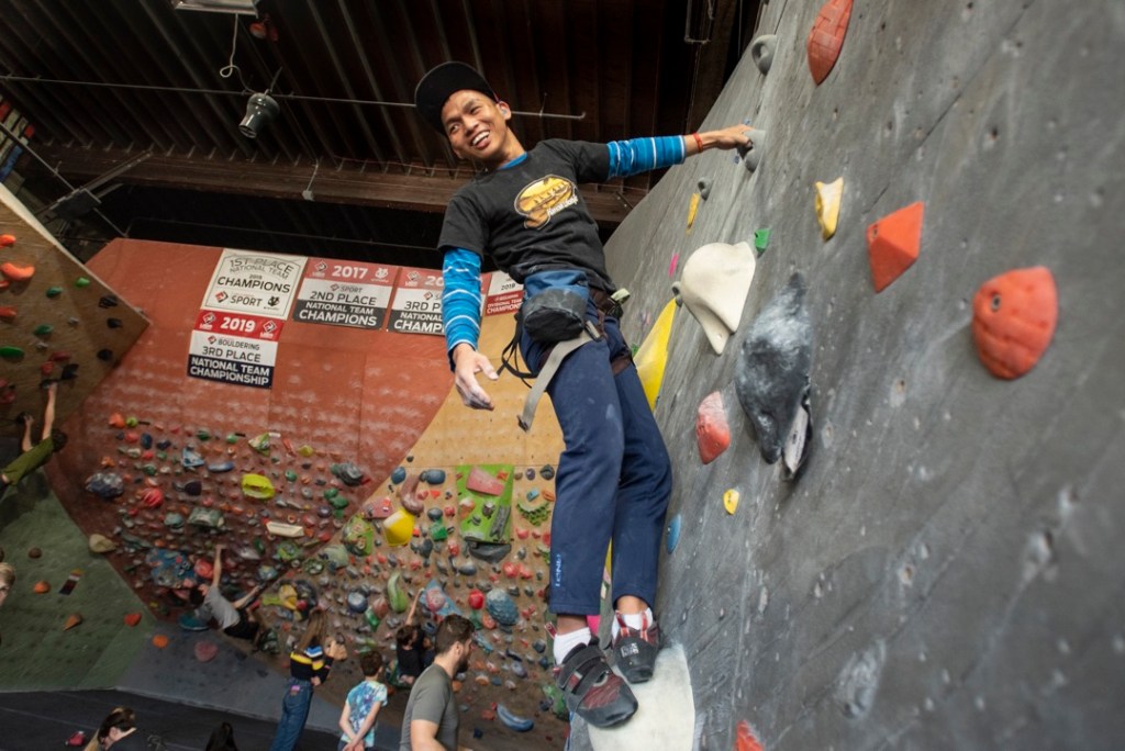 Thein Mana takes a rest, hanging from his left hand and standing on a large hold on an indoor bouldering wall.