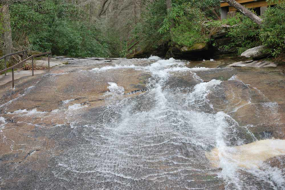 A mellow whitewater rapid smoothes its way over rocks with green trees in the background