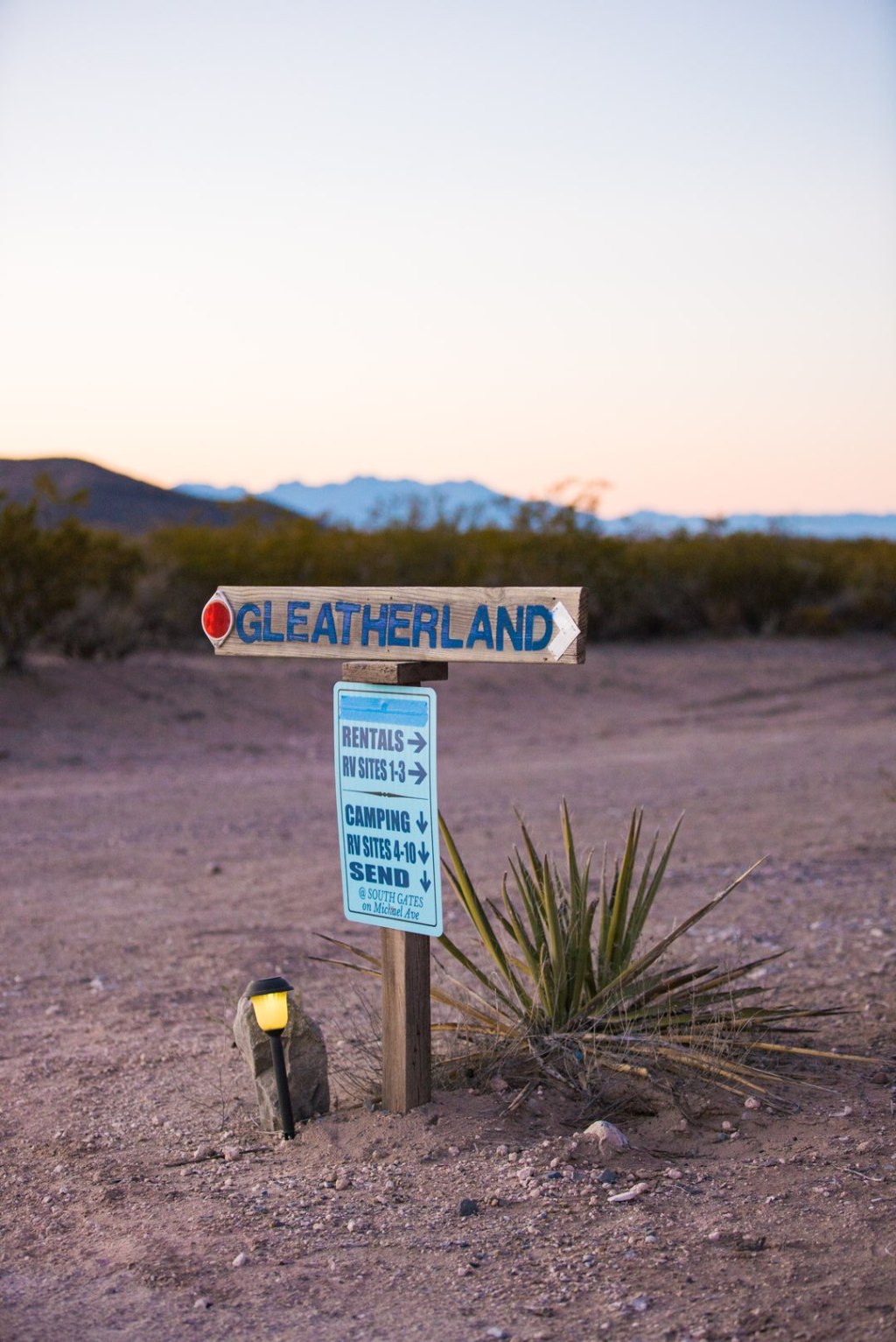 A sign along a dirt road points to a campsite, with a sunset over rocks on the horizon.