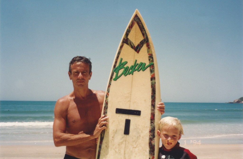 A young boy stands beside his father with a very tall surfboard with the beach in the background