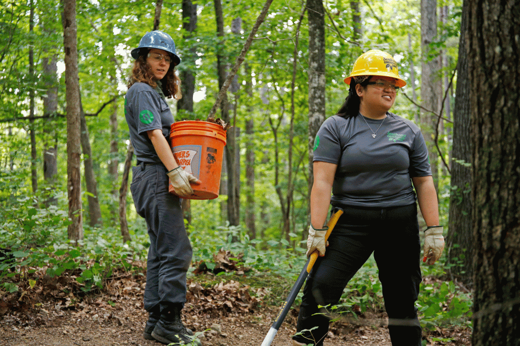 A woman pauses for a break while wearing a hard hard, while another looks on with a utility bucket