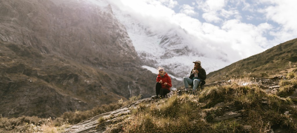 Two hikers sit on the trail with a backdrop of a snowy mountain.