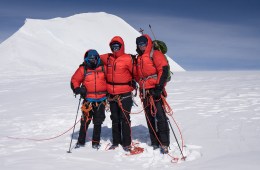 Three climbers in red puffy jackets stand, arms linked, tied into a rope on a snowy mountain.