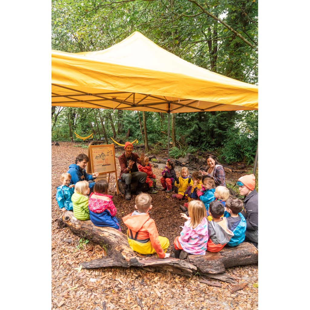 Children sit under a tent for an outdoor preschool lesson