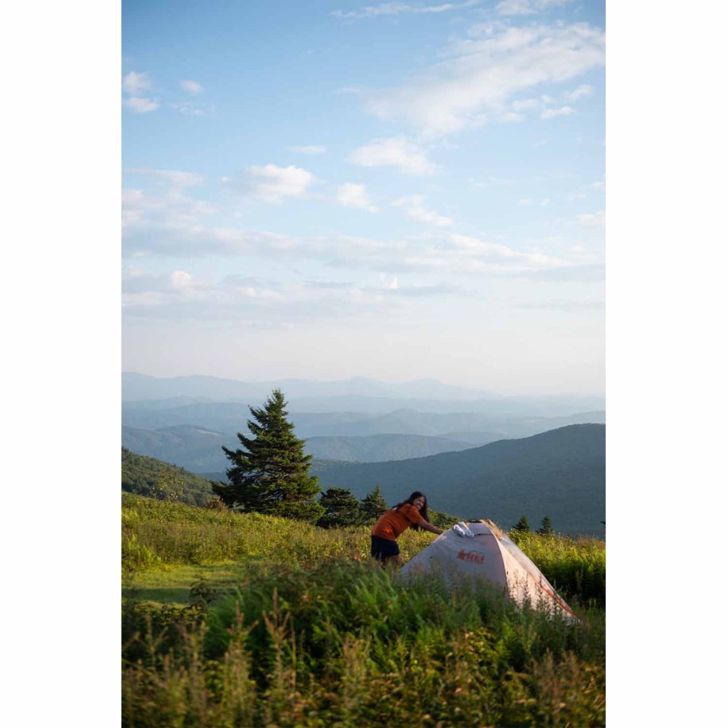 A woman sets up an REI tent in a field with a soft blue sky in the background