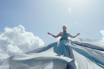 Pattie Gonia stands with a background of sky, flowing dress coming towards the camera.