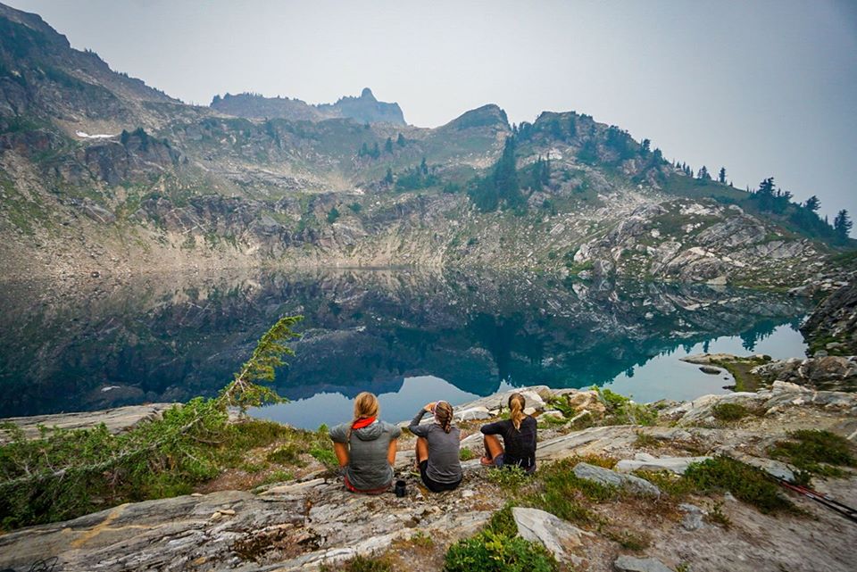Three friends sit beside a blue-green lake in the mountains