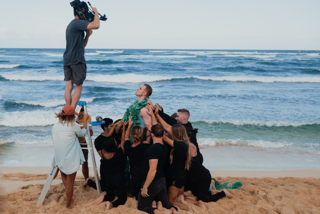 A videographer sands on a ladder, filming Pattie Gonia surrounded by cast members.