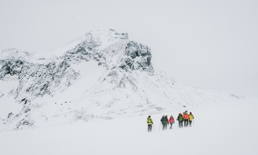 A group of people in colorful outerwear hike beside a large snow-covered mountain.