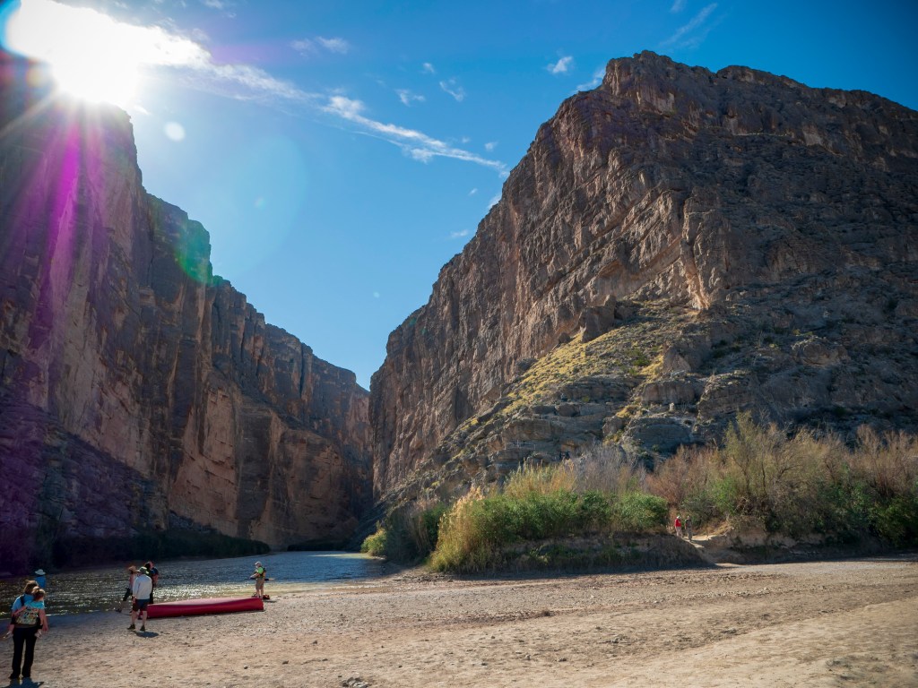 People standing on a riverbank next to canoes and a river below canyon walls during the daytime.