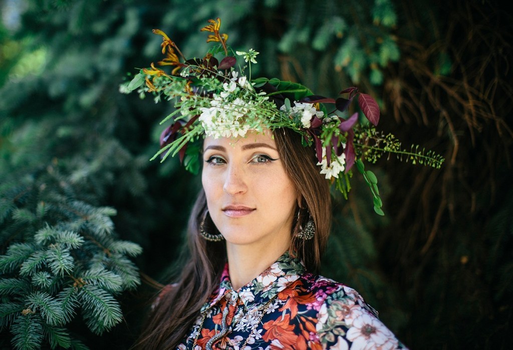 Jenny Kendler poses with a flower crown in front of green foliage.