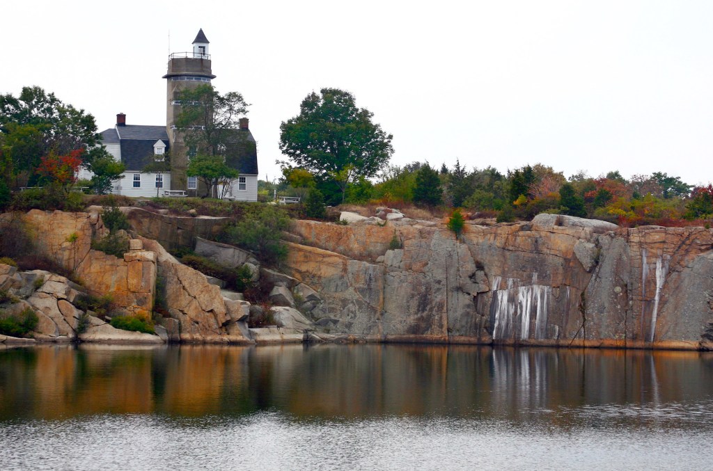 A still body of water is in the foreground with a cliff that has a lighthouse and some plants on top against a cloudy sky.