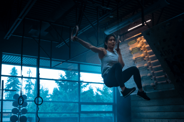 Alannah Yip training on rings inside a dark climbing gym backlit by blue lighting.