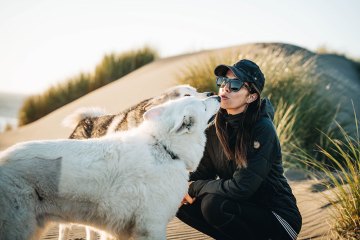 Ally Coucke is kneeling on the beach with her two dogs kissing her face.