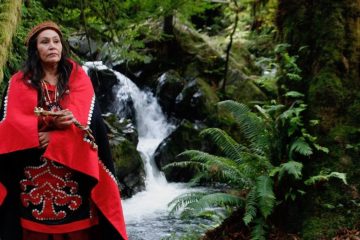 Harvest Moon, a Quinault native elder and storyteller, stands in front of a waterfall and ferns wearing a red cloak.