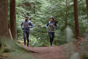 A couple jogs through the forest together surrounded by tall evergreen trees