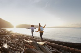 Two people walking on logs on a beach with sun setting.