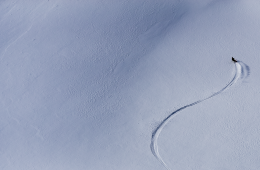 Michelle Parker captured from an aerial camera skiing down a mountain. She is a tiny bright spot in a sea of white.