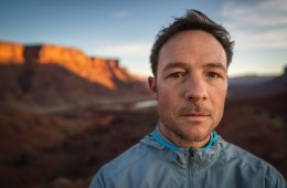 Adam Campbell is staring at the camera with a mountain range in the background, lit up by the sun.