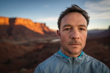 Adam Campbell is staring at the camera with a mountain range in the background, lit up by the sun.