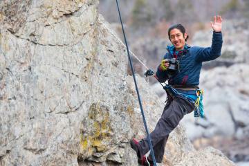 Smiling rock climber hanging from fix line holding camera and waving.
