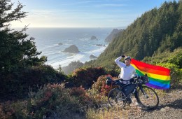 Mikah Meyer is standing on a trail in front of his bike holding a pride flag above his head with a background of trees and the ocean.