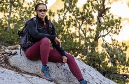 Nicole Snell, wearing purple pants, hiking boots and a black jacket, is sitting on a rock and looking at the camera with trees in the background.