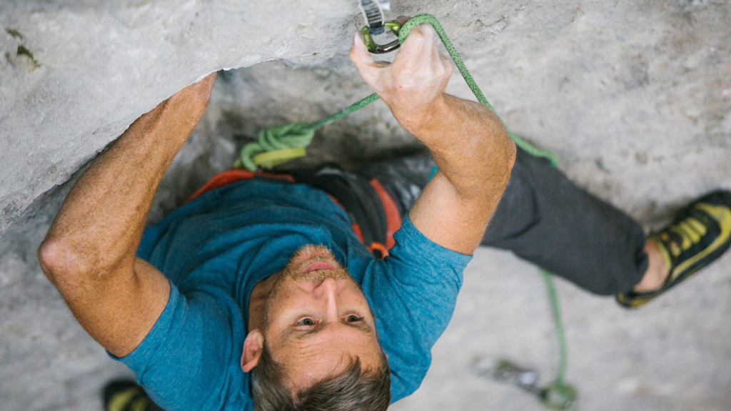 A professional rock climber clips a green climbing rope while balancing on the rock wall.
