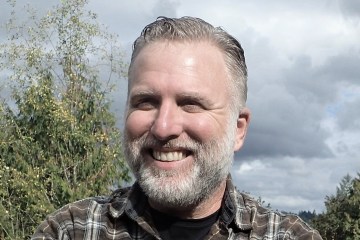 A close up shot of Cliff Barackman's face as he smiles at the camera. He's wearing a plaid shirt and has grey hair and a grey beard.