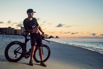 Ryan Kinder dressed in a bike kit is leaning against a bike that's on the sand on a white sand beach. He's looking out at the ocean and the sky is orange and blue from a sunset.