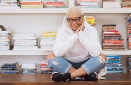 Angela Medlin is sitting on a table in a white hoodie and jeans while smiling at the camera.