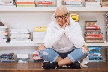 Angela Medlin is sitting on a table in a white hoodie and jeans while smiling at the camera.