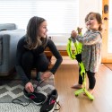 A woman with brown hair kneels down to lace her running shoes, while smiling at her toddler-aged daughter, who is next to her, holding a reflective running vest and also smiling.