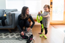 A woman with brown hair kneels down to lace her running shoes, while smiling at her toddler-aged daughter, who is next to her, holding a reflective running vest and also smiling.