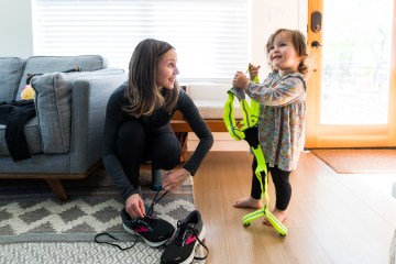 A woman with brown hair kneels down to lace her running shoes, while smiling at her toddler-aged daughter, who is next to her, holding a reflective running vest and also smiling.