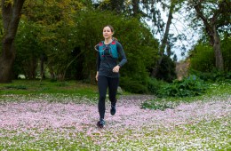 Woman with brown hair in a ponytail run across a field covered in pink flower petals. She's wearing a red and blue hydration vest and black athletic clothing.