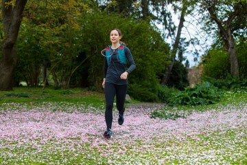 Woman with brown hair in a ponytail run across a field covered in pink flower petals. She's wearing a red and blue hydration vest and black athletic clothing.