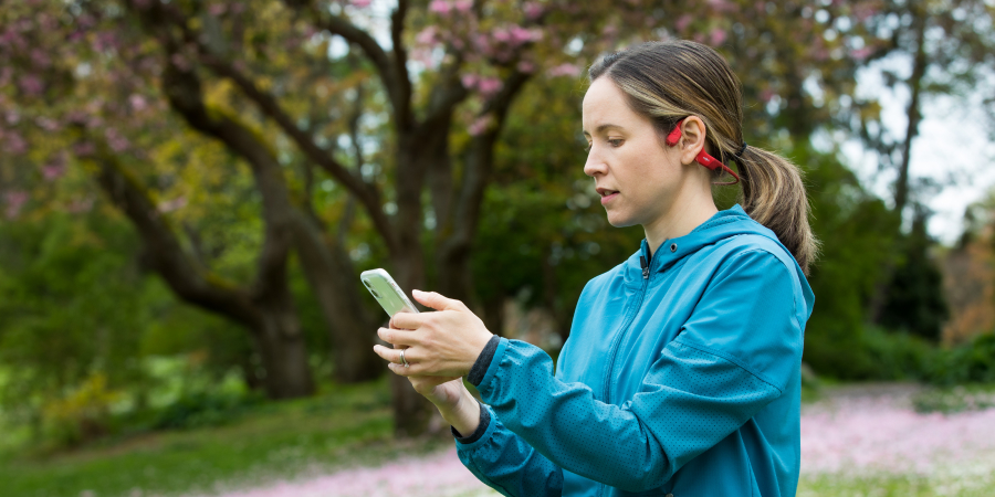 Woman with brown hair in a ponytail, wearing a bright blue running rain jacket and red open-ear running headphones, looks at her phone. 